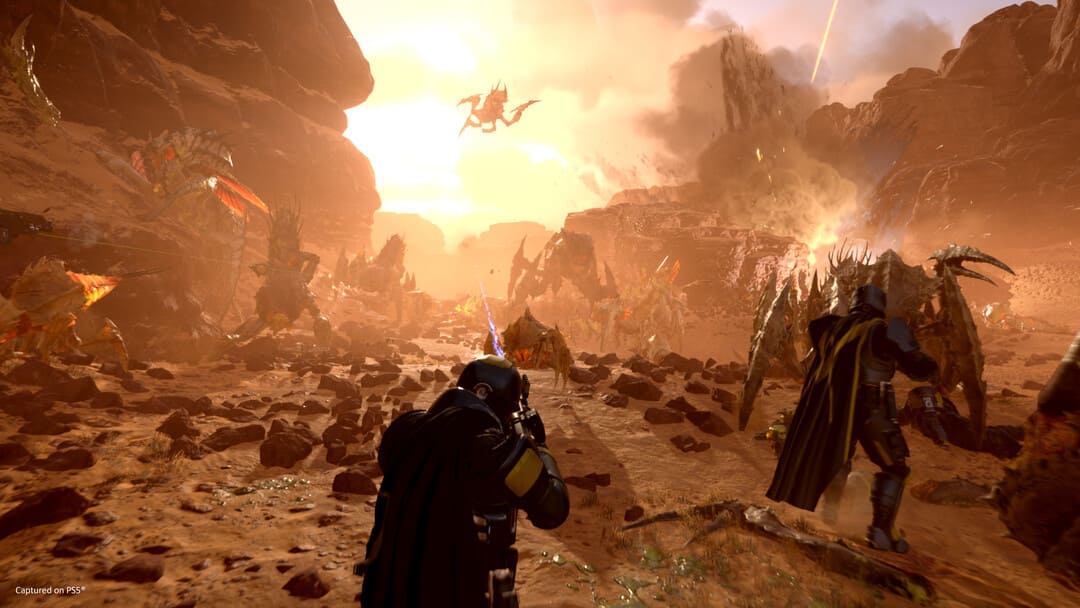 ゲームHelldivers2のスクリーンショットは、人間と昆虫の間の火星のような惑星への攻撃を示し、背景に爆​​発があります