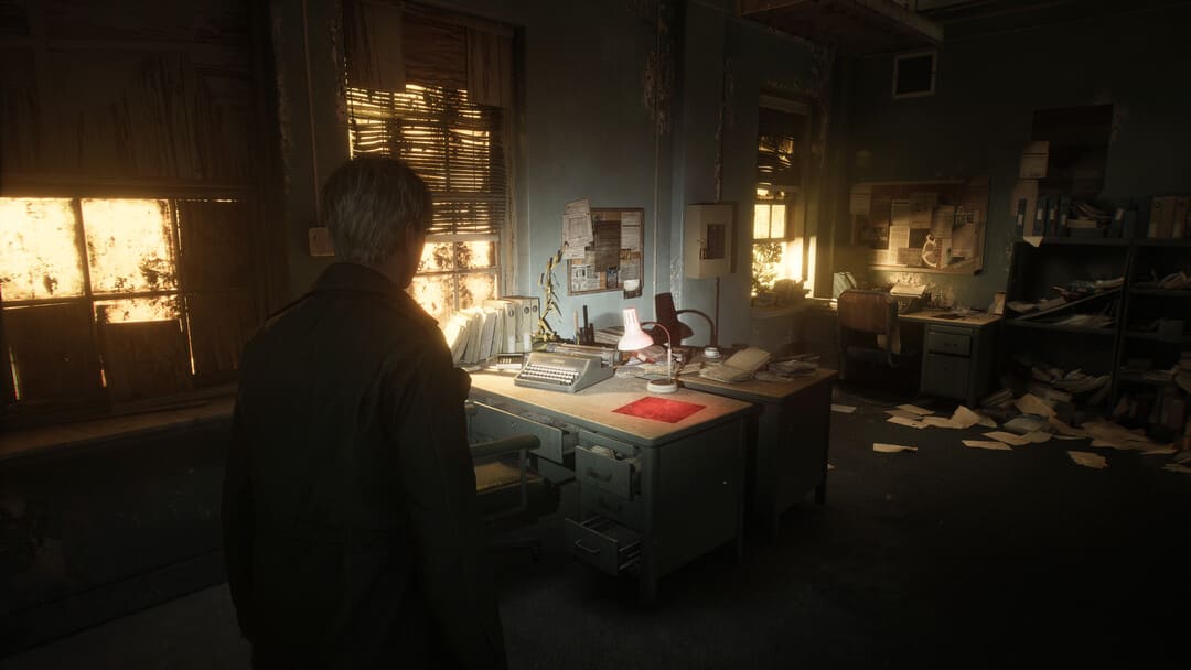 ゲームサイレントヒル2（2024）のスクリーンショットは、どこにでもタイプライターとルーズペーパーを備えた放棄されたオフィスの中の男性を示しています